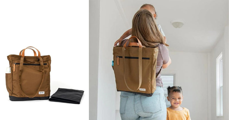 Parent carrying the Tote Pack Baby diaper bag as a backpack while holding a baby, with a toddler smiling in the background, emphasizing its practicality and family-friendly design.