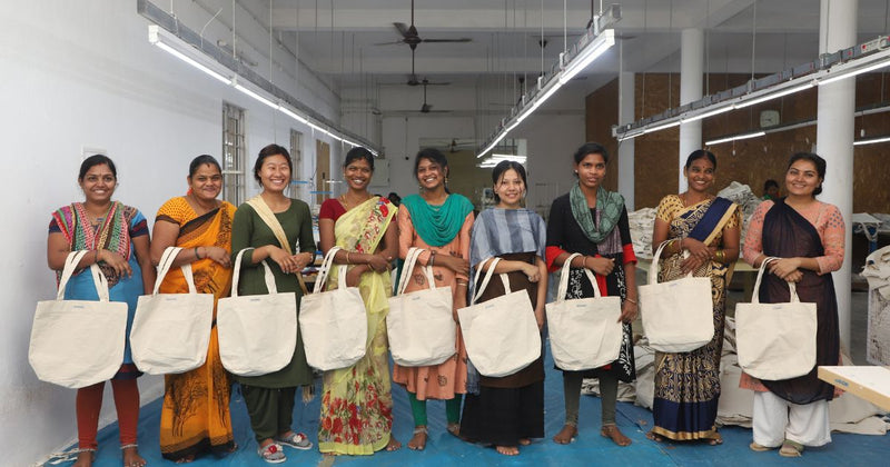 A group of eight women in traditional South Asian attire, smiling and standing in a workshop, each holding a canvas tote bag. Bright lighting overhead.