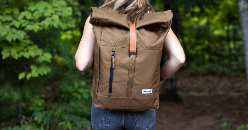 A person wearing a tan roll-top backpack from MADE FREE with a leather strap and zipper pocket, standing in a lush green outdoor setting