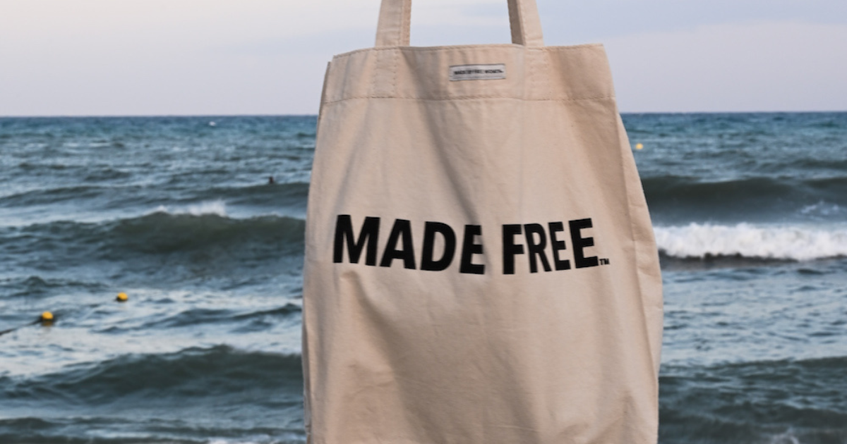  A beige market canvas tote bag with bold black text that reads "MADE FREE" is pictured against the backdrop of ocean waves.