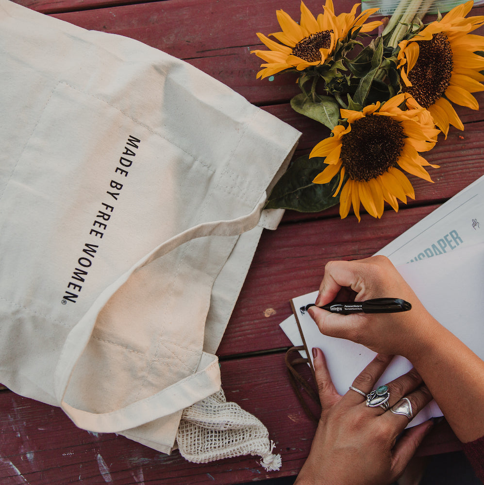 A cream-colored canvas bag with the text "MADE BY FREE WOMEN" placed next to a bouquet of sunflowers and a hand writing in a notebook.