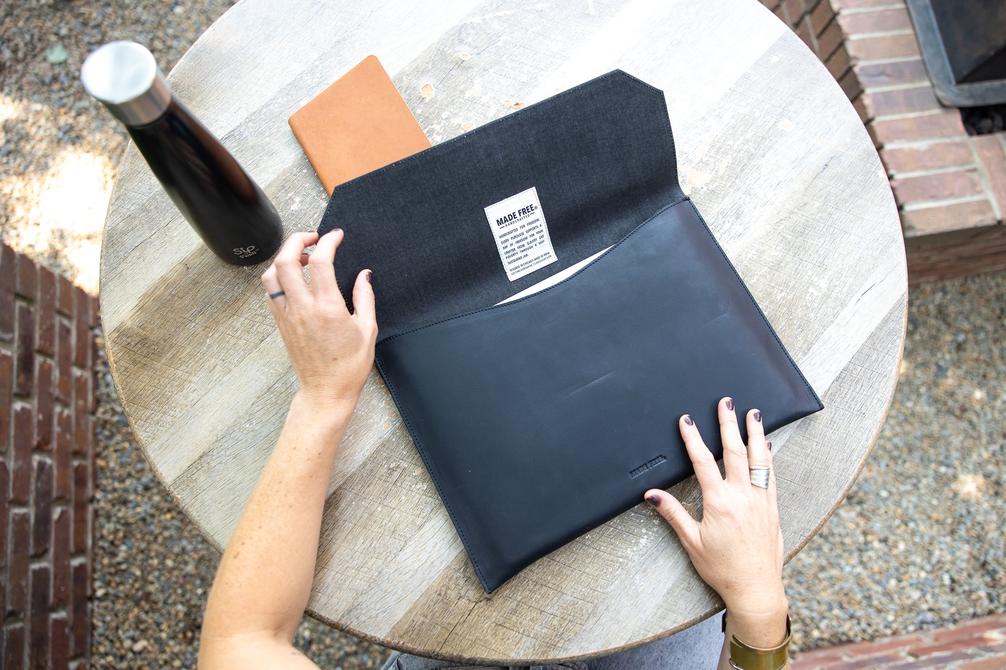 A person opening a black leather laptop sleeve on a round wooden table with a black water bottle and orange notebook beside it.