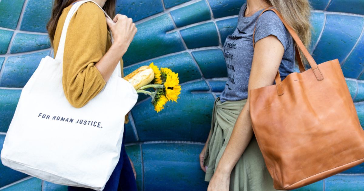 Two women walking side by side, each carrying eco-friendly and ethically-made tote bags. The woman on the left holds a canvas tote bag that reads "For Human Justice" while the woman on the right carries a tan leather tote. The canvas bag contains sunflowers and a fresh baguette. These sustainable, fair trade bags promote ethical fashion, aligning with environmentally-conscious choices and the fight against human trafficking. Perfect for an eco-friendly lifestyle, these tote bags embody a blend of style and 