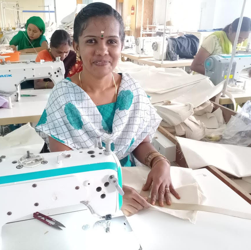 A woman wearing a sari sits at a sewing machine in a bright workshop, smiling at the camera.