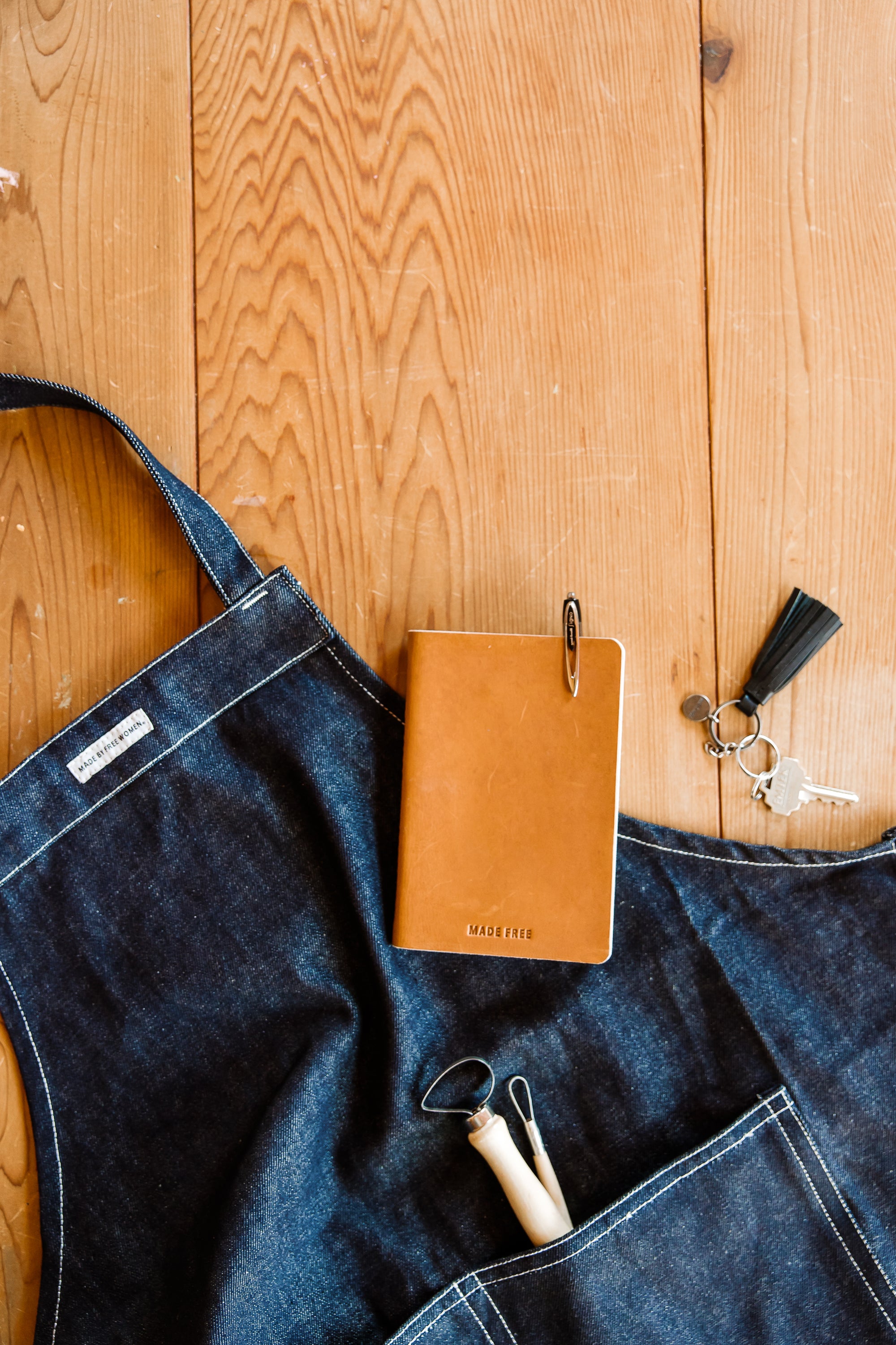 A flat lay of a denim apron with a brown leather notebook, keys, and a small pottery tool on a wooden table.