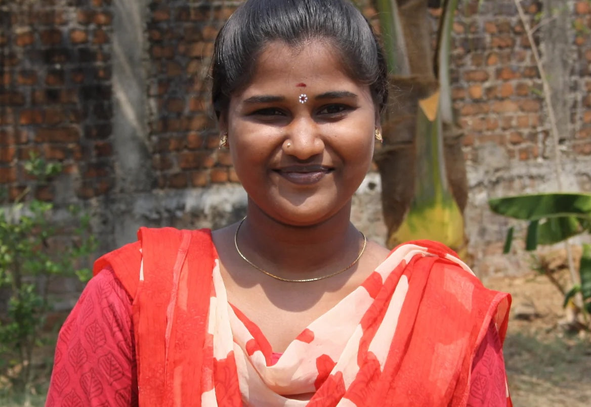 A young woman in a red sari smiles at the camera outdoors, standing against a brick background with greenery behind her.