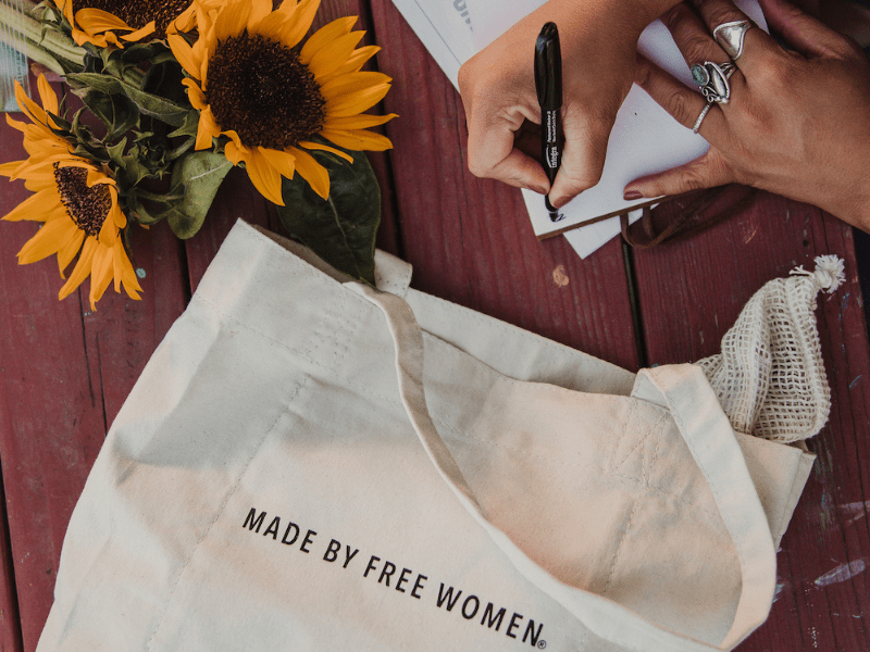 Canvas tote with "Made by Free Women" text, farmers market, sunflowers, and hands writing in a notebook, symbolizing ethical fashion and women's empowerment.
