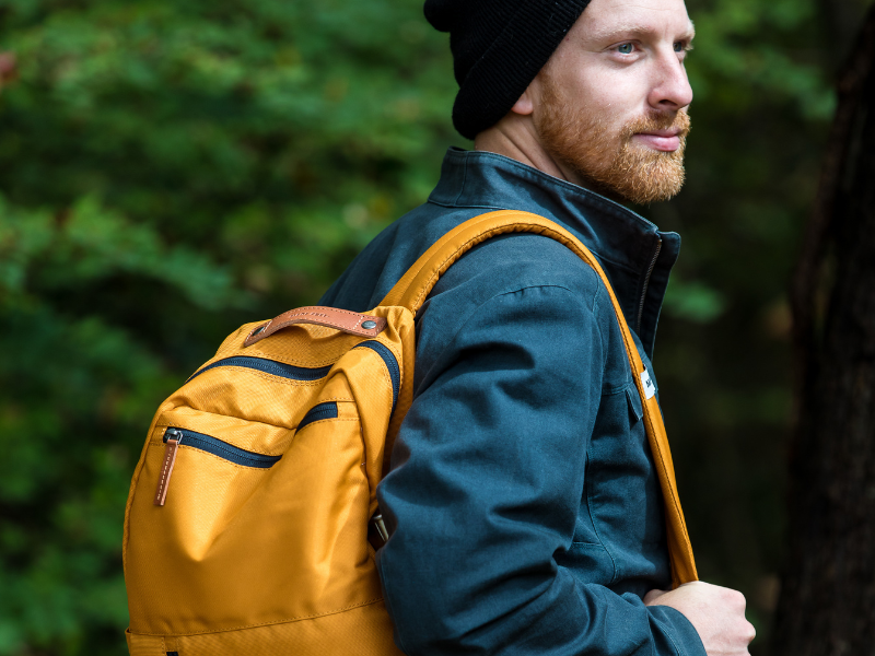 A man wearing a black beanie and blue jacket carries a mustard yellow recycled polyester backpack in a forest, highlighting eco-friendly and sustainable fashion.