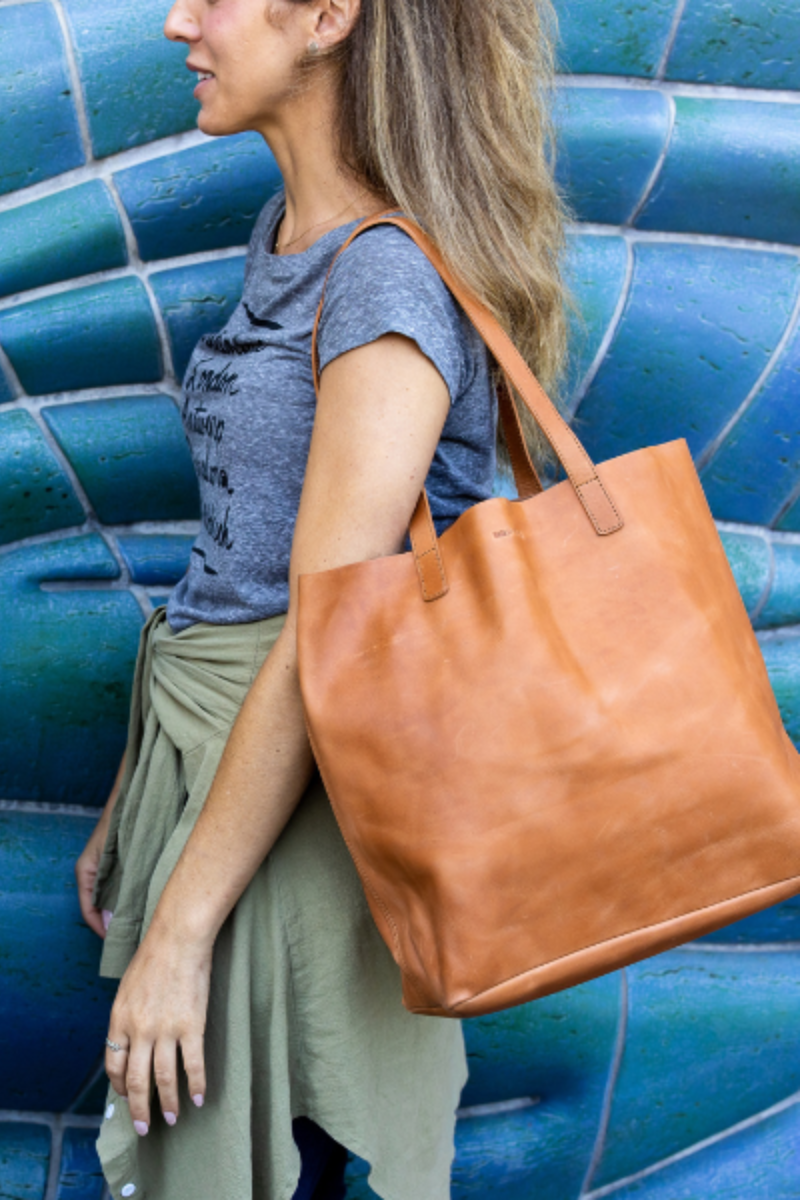 A woman carries a large veg tanned leather tote bag in color carmel, wearing a casual outfit of a grey tee and green wrap, standing in front of a blue mosaic wall. The tote reflects minimalist and sustainable style.